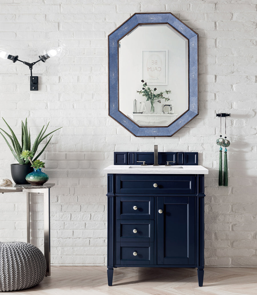 A Brittany 30" Vanity with a white countertop stands against a white brick wall, beneath a blue-edged octagonal mirror. Accented by North American Birch and decor, this setup creates a modern, minimalist bathroom look.