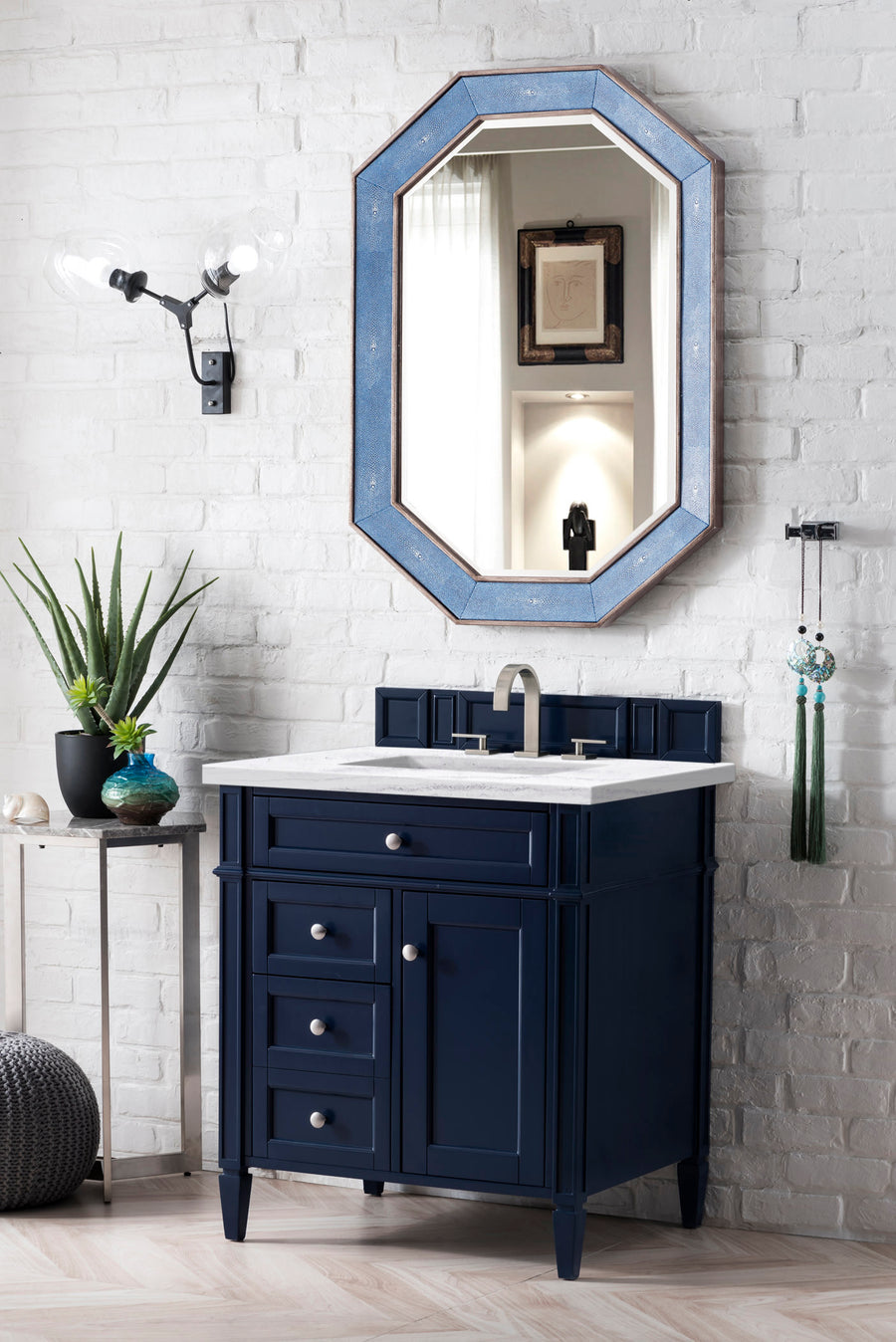 A modern bathroom features a blue vanity with silver hardware, a white countertop, a rectangular sink, a tall octagonal mirror above, and a brick wall background decorated with a plant and two light fixtures.