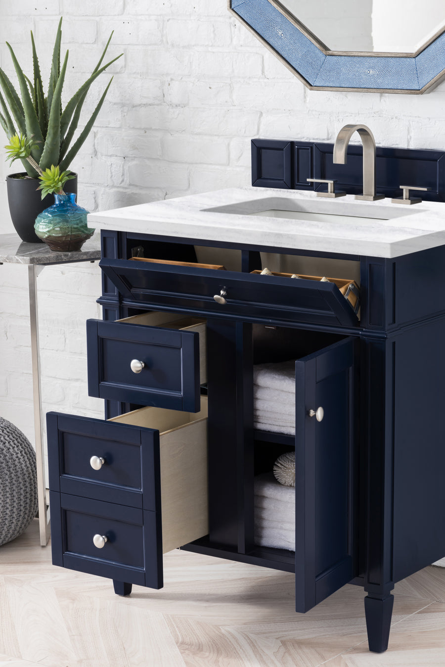 A navy blue bathroom vanity with a marble top, open drawers and cabinet doors showing towels inside, sits against a white brick wall. A modern faucet and octagonal mirror are above, and a small table with plants stands nearby.