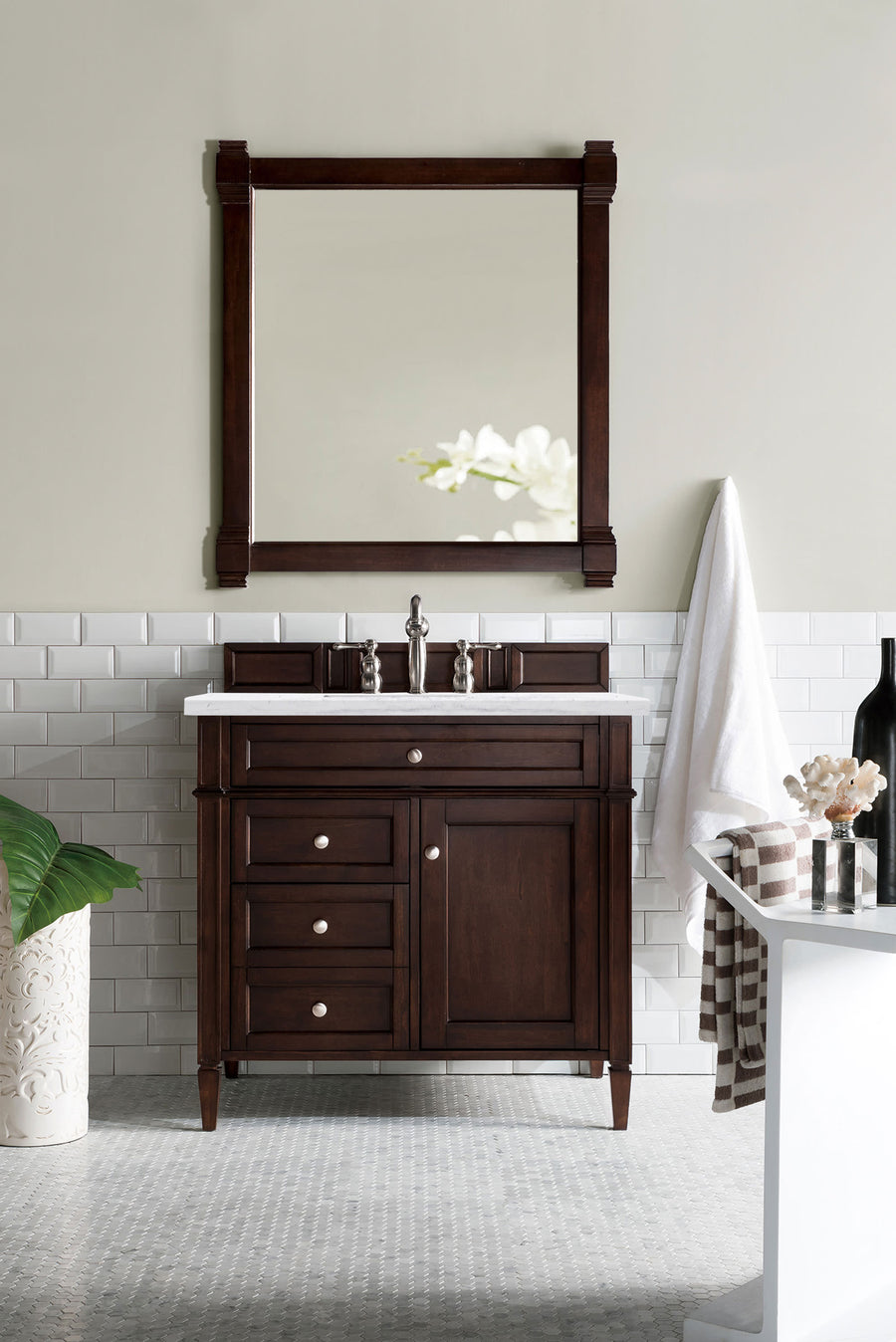A bathroom features the Brittany 36" Vanity with a white countertop and silver faucet under a matching framed mirror. White subway tiles line the lower wall, with decor like a plant, vases, towels, and a nearby bathtub.
