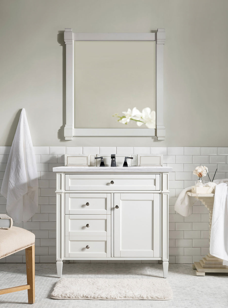 The Brittany 36" Vanity with a marble top and matching mirror is accented by white towels and a reflected flower, while white subway tiles and light decor enhance the clean, minimalist look of this hardwood cabinet.