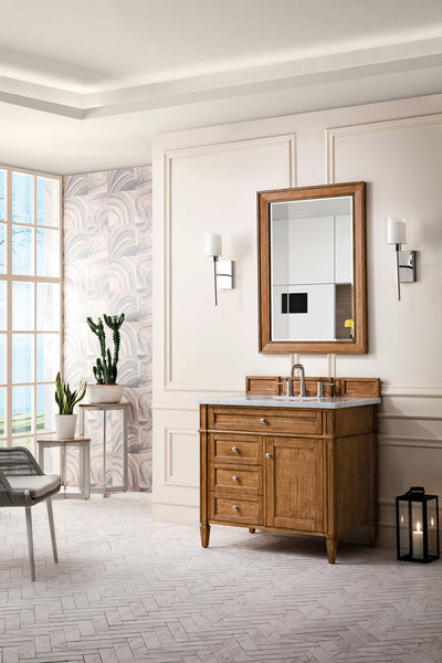 A stylish bathroom with a wooden vanity, rectangular mirror, and wall sconces. There is a window with natural light, potted plants, a lantern on the floor, and patterned wallpaper on one wall.