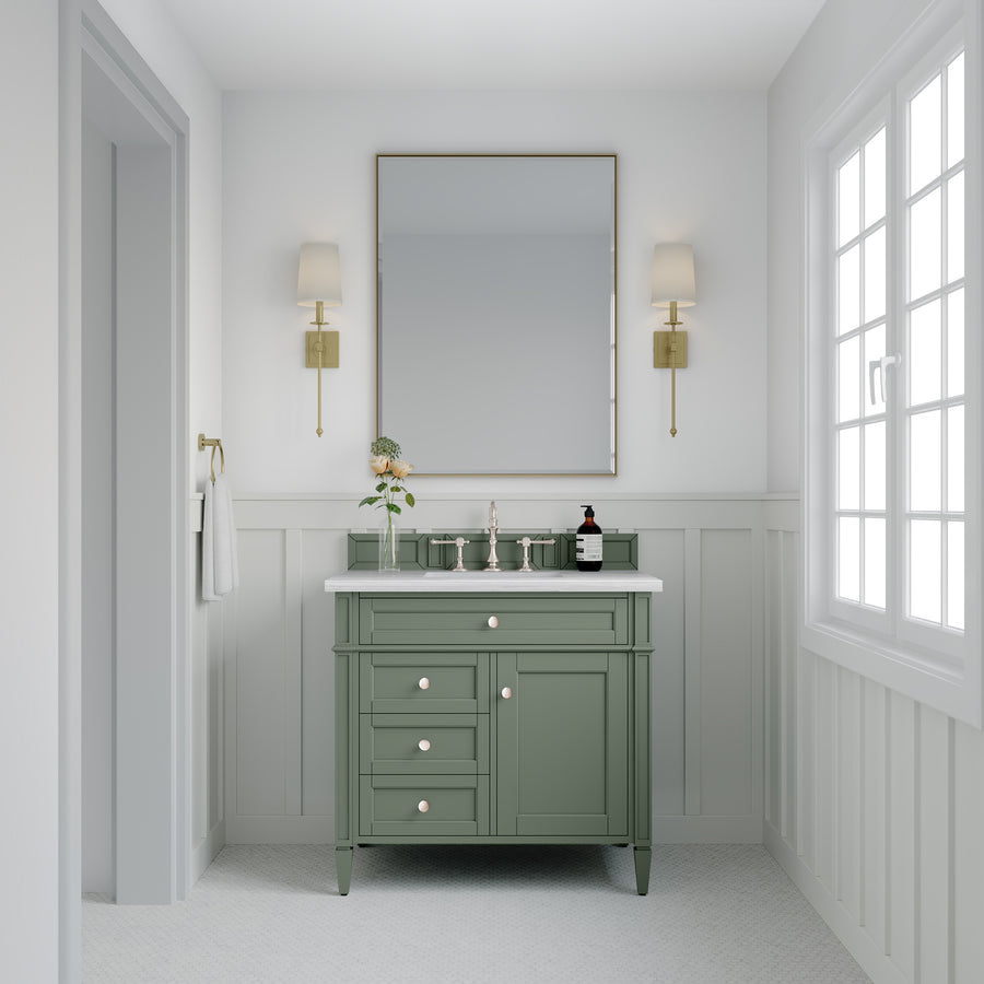 A bright bathroom features a green Brittany 36" Vanity with double faucets, a large framed mirror, brass sconces, a hand towel to the left, a plant on the counter, and sunlight from big right-side windows.