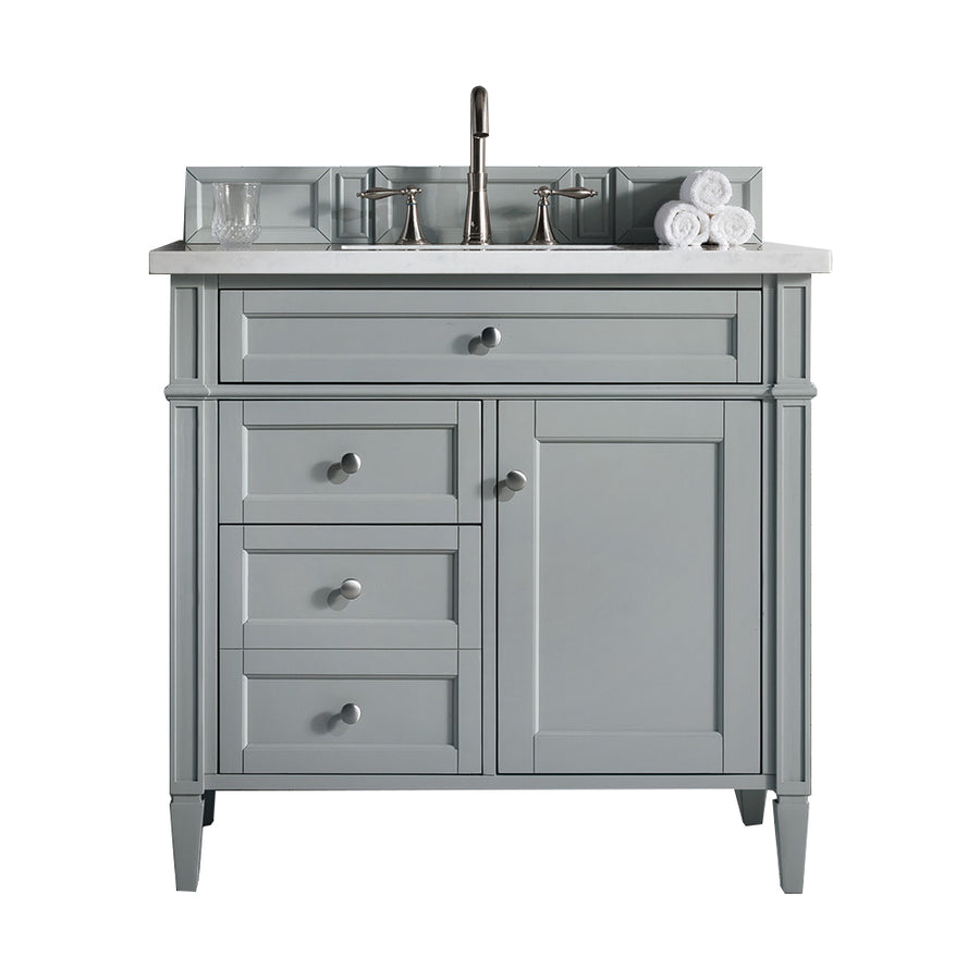 A light gray bathroom vanity with a marble countertop, silver faucet, three stacked drawers, a cabinet, and a backsplash. Rolled white towels and a glass sit on the counter.
