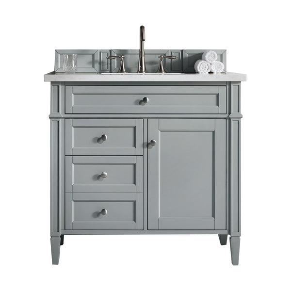 A light gray bathroom vanity with a marble countertop, silver faucet, three stacked drawers, a cabinet, and a backsplash. Rolled white towels and a glass sit on the counter.