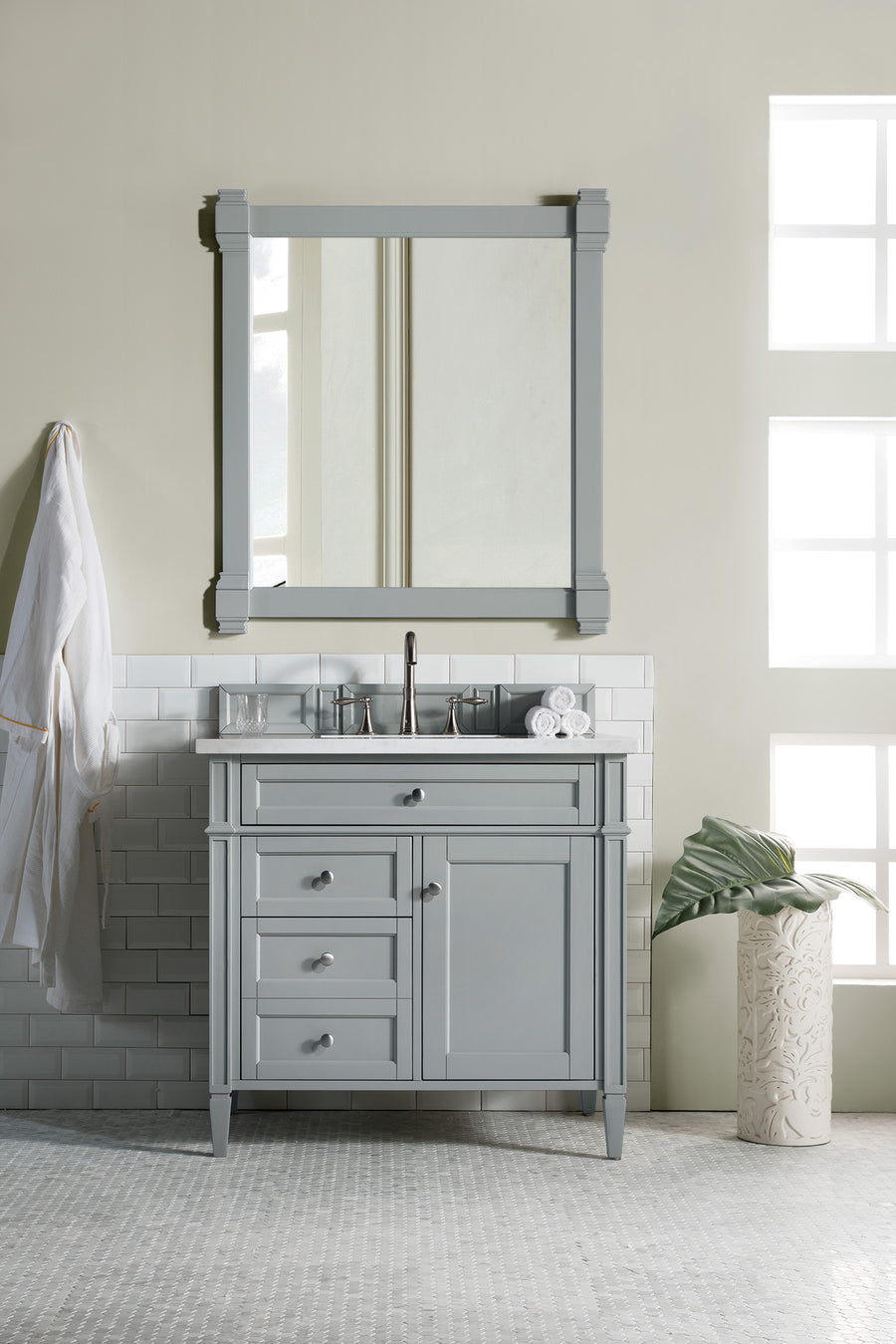 A Brittany 36" Vanity in light gray with a matching mirror, silver faucet, and drawers stands by a tiled wall. A robe hangs to the left while a vase with a large leaf sits to the right near bright windows.