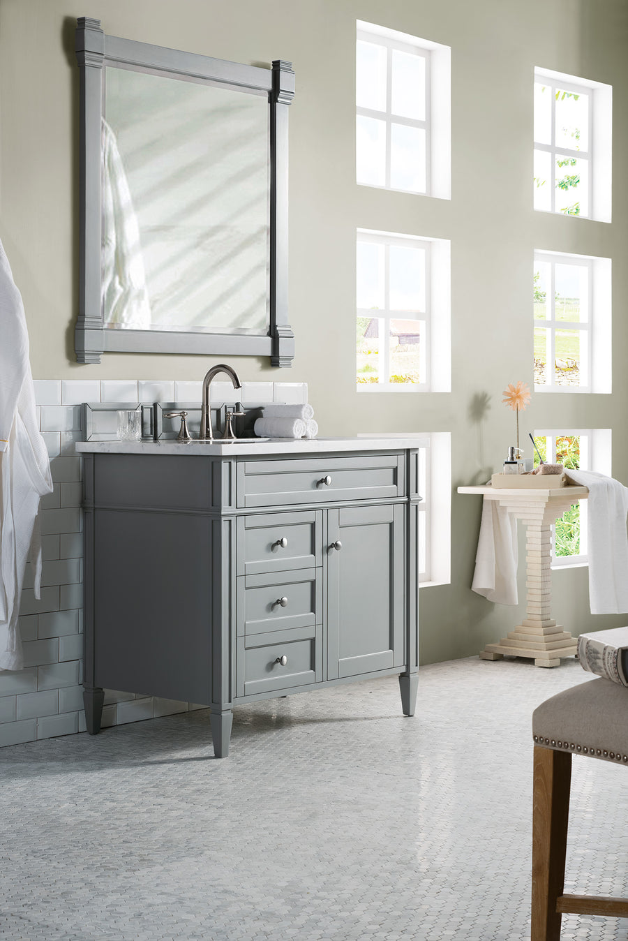 A modern bathroom with a gray vanity, silver faucet, large framed mirror, and light green walls. Four windows let in natural light, and a small table holds a flower in a vase and folded towels. White tile floor and backsplash.