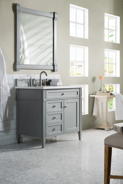A modern bathroom with a gray vanity, silver faucet, large framed mirror, and light green walls. Four windows let in natural light, and a small table holds a flower in a vase and folded towels. White tile floor and backsplash.