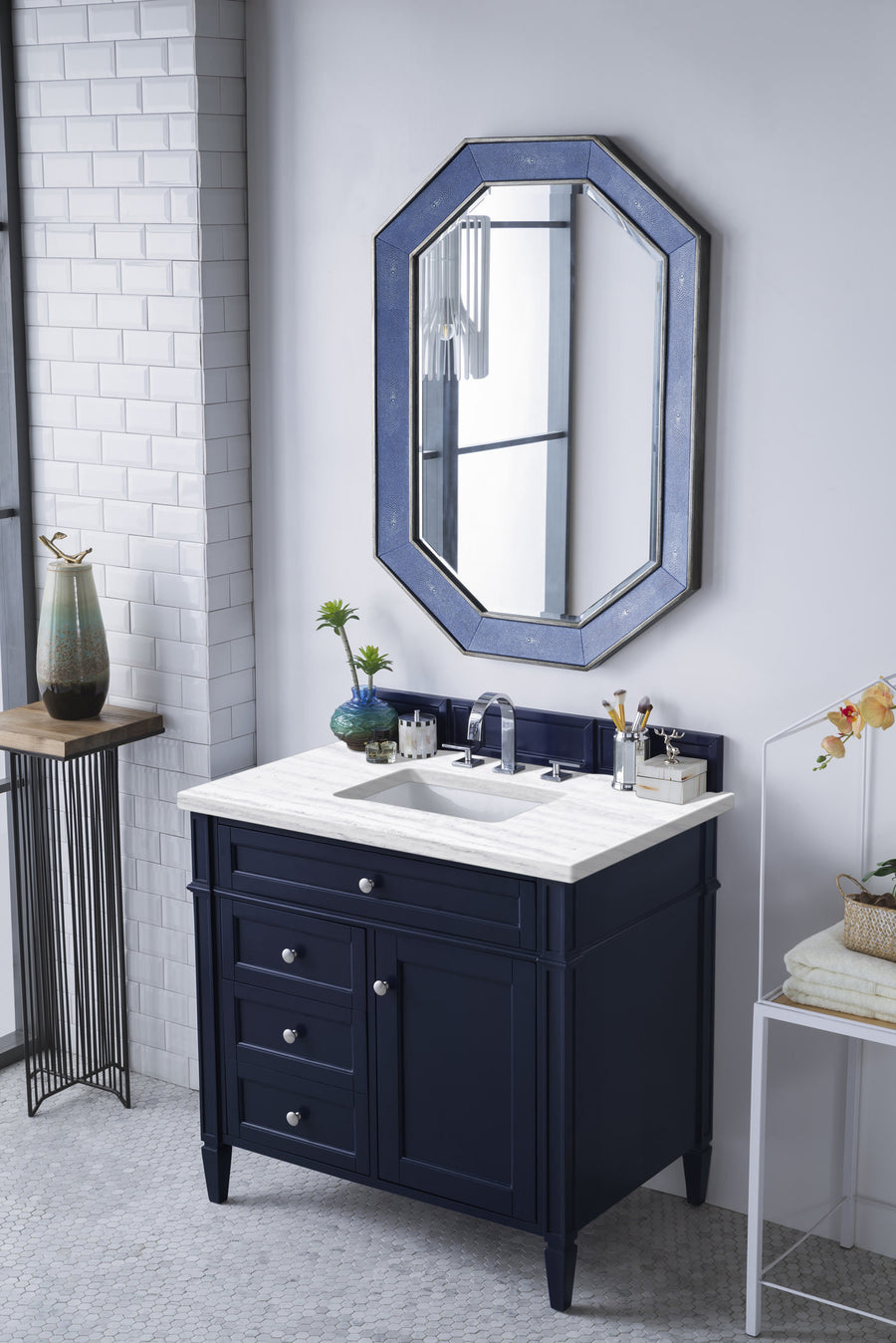 A modern bathroom with a navy blue vanity, white countertop, silver fixtures, an octagonal mirror above, white tile walls, and decorative items including vases, towels, and a small plant.