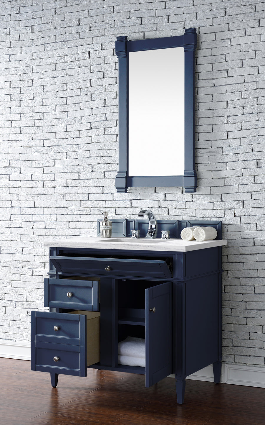 A navy blue bathroom vanity with drawers and doors open, topped with a white countertop, chrome faucet, soap dispensers, and rolled towels. A matching framed mirror hangs on a white brick wall above dark wood flooring.