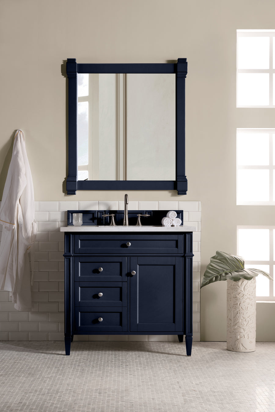 A bathroom featuring the Brittany 36" Vanity with matching framed mirror, white sink, silver faucet, folded towels, a white robe on the left wall, and a plant in a white vase on the right. Natural light fills the room.
