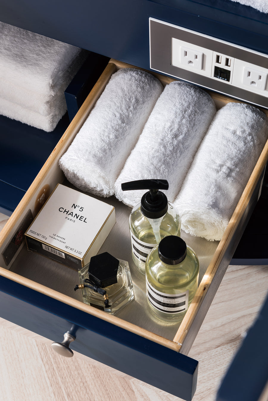 A bathroom drawer neatly organized with three rolled white towels, a box of Chanel No. 5 soap, a perfume bottle, and two pump bottles of liquid soap. An electrical outlet panel is visible above the drawer.