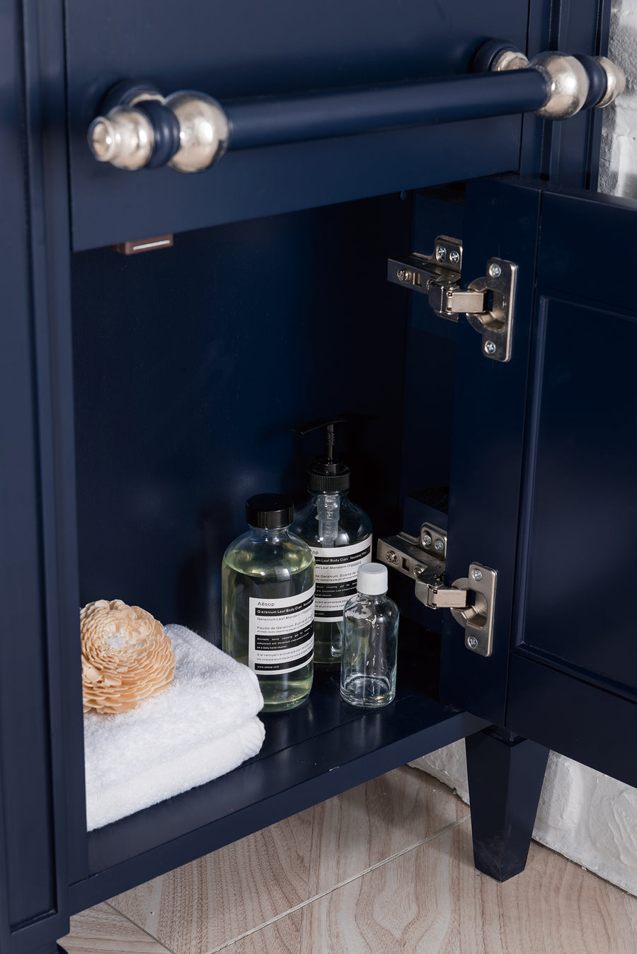 A navy blue bathroom cabinet with an open door reveals a white folded towel, a natural sponge, and several bottles of liquid soap and toiletries on a shelf inside. The cabinet stands on a tiled floor.