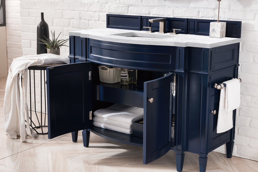 A navy blue bathroom vanity with open cabinet doors shows neatly folded white towels and storage baskets inside, topped with a white countertop and sink. A round side table holds decor and a draped towel. White brick wall background.