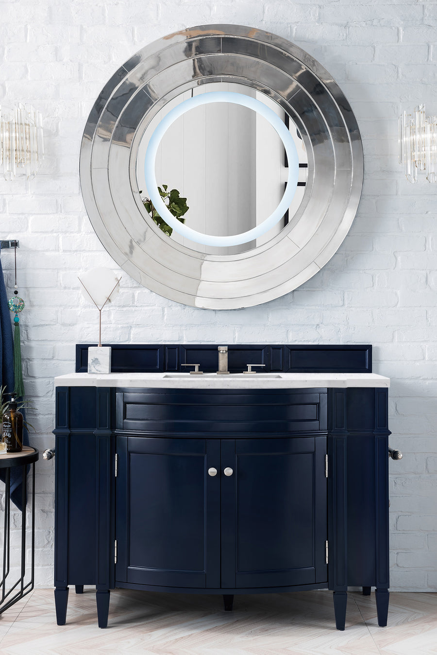 The Brittany 46" Vanity is a modern dark blue hardwood cabinet with a marble countertop and double faucet, set beneath a round silver-framed mirror on a white brick wall accented by decorative lights.