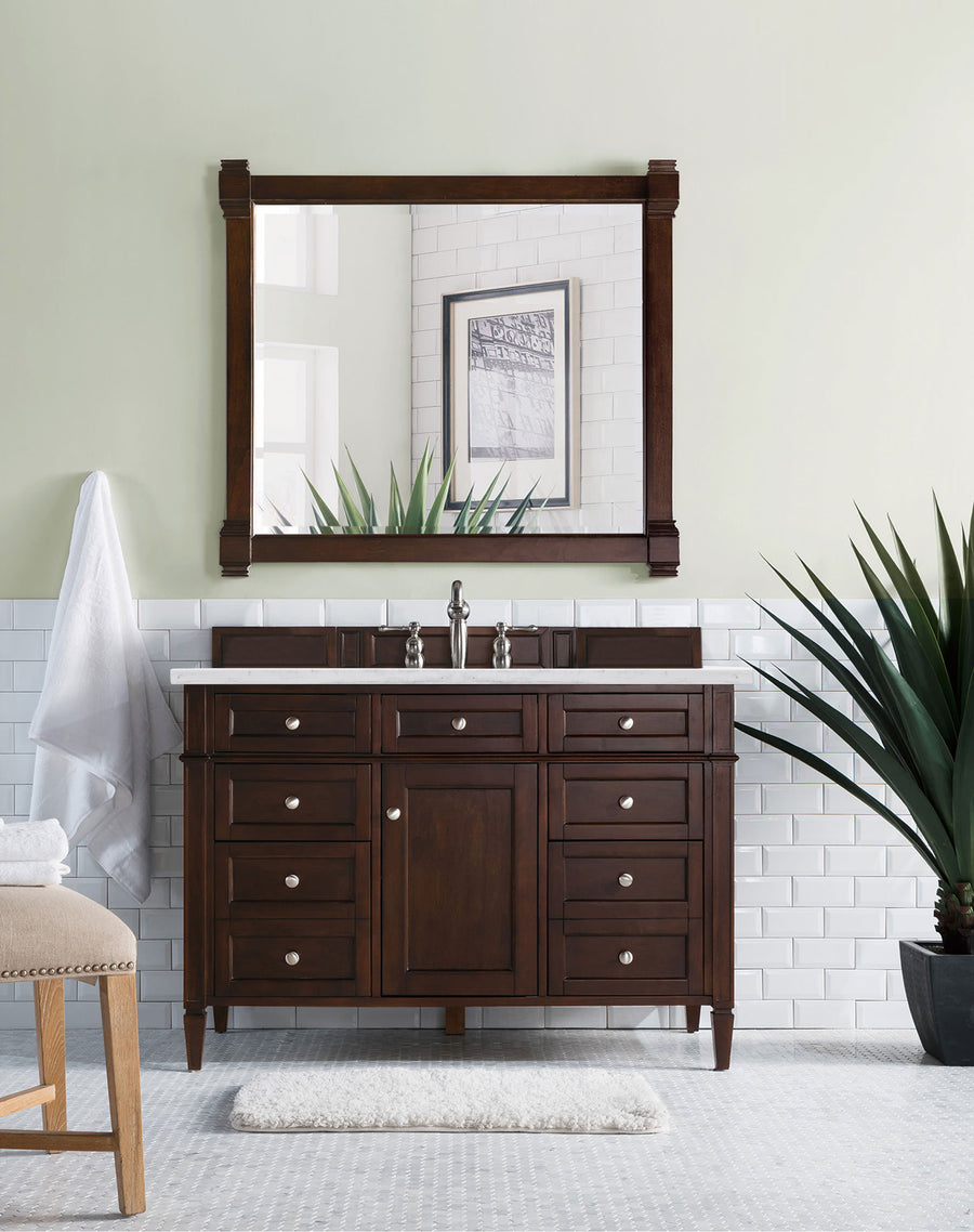 A bathroom with the Brittany 48" Vanity double sink, wall mirror, white subway tile, potted plant, towel on a hook, and beige stool with folded towels against pale green walls—luxurious bathroom style.