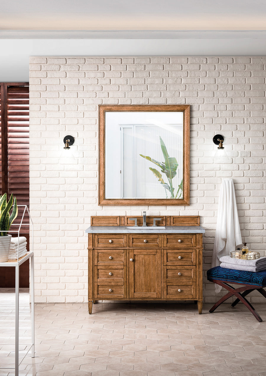 A bathroom with white brick walls features a Brittany 48" Vanity with drawers, a large framed mirror, two wall lights, a blue cushioned stool, a towel, and potted plants. A window reveals green leaves outside.