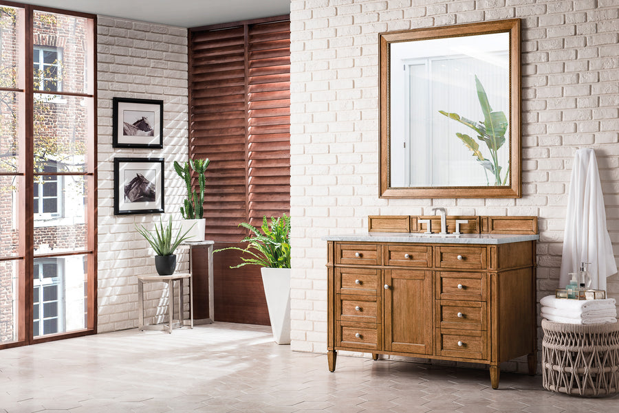 A bright, modern bathroom with white brick walls, a wooden vanity with a mirror, potted plants, wall art, and large windows allowing natural light to fill the space. Towels are stacked in a basket beside the vanity.