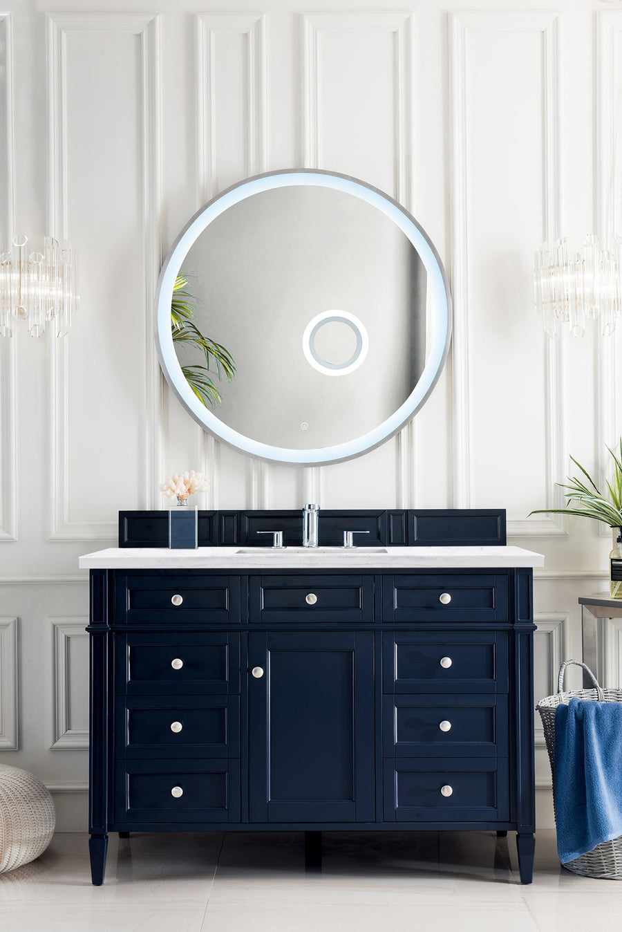 A modern bathroom featuring white paneled walls, a round backlit mirror, the Brittany 48" Vanity with silver hardware and marble top, luxury furniture accents, and decorative plants on each side.