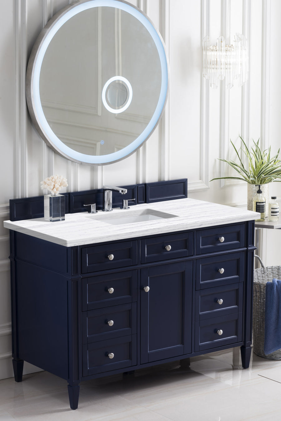 A modern bathroom features a navy blue vanity with a white countertop, silver hardware, a rectangular sink, and a large round mirror with a built-in LED light on a white paneled wall. Decorative items and towels are nearby.