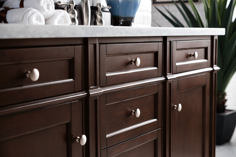 A close-up of dark wood bathroom cabinets with round cream knobs, under a white countertop. Rolled white towels, silver accessories, and a blue vase are visible on the counter; a green plant is in the background.