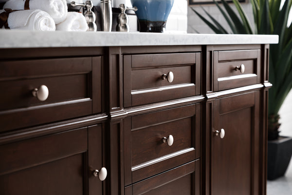 A close-up of dark wood bathroom cabinets with round cream knobs, under a white countertop. Rolled white towels, silver accessories, and a blue vase are visible on the counter; a green plant is in the background.