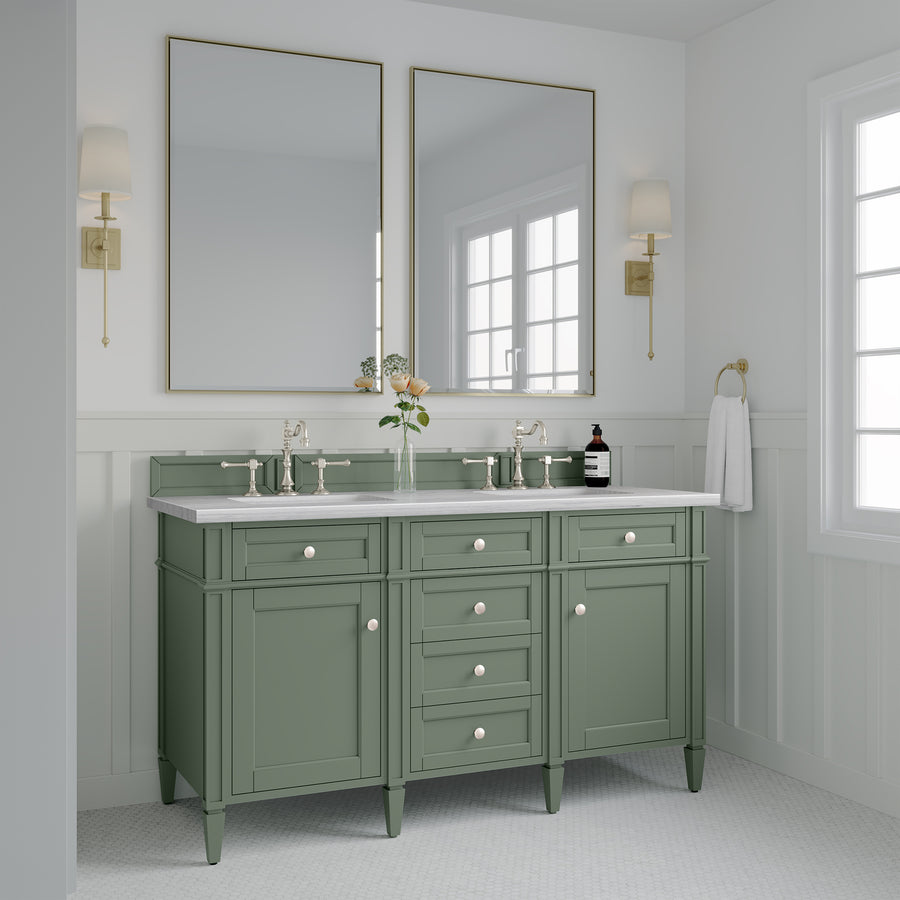 A bathroom with a green double-sink vanity, two rectangular mirrors, wall sconces, a white countertop, and white paneled walls. A vase with flowers and toiletries are on the vanity. Sunlight streams in through a window.