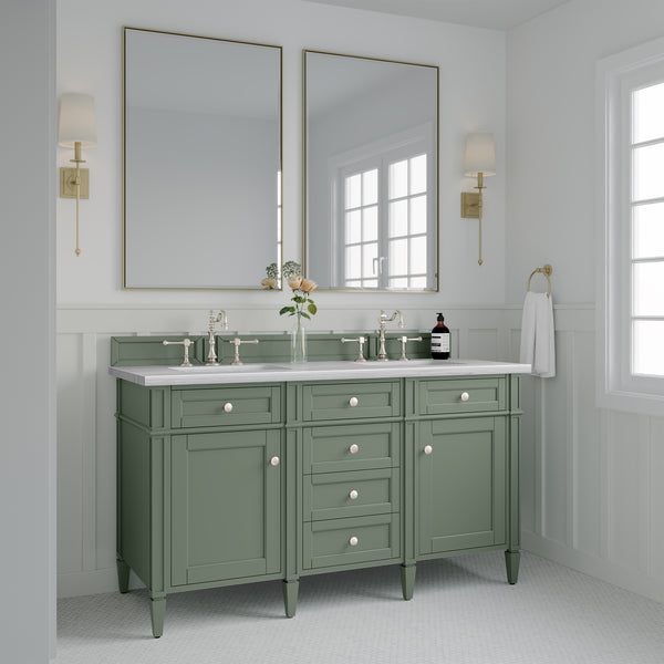 A bathroom with a green double-sink vanity, two rectangular mirrors, wall sconces, a white countertop, and white paneled walls. A vase with flowers and toiletries are on the vanity. Sunlight streams in through a window.