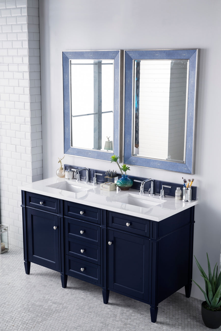 A modern bathroom features a navy blue double-sink vanity with silver hardware, a white countertop, and two rectangular mirrors with blue frames above. The vanity is neatly arranged with toiletries and decorative plants.