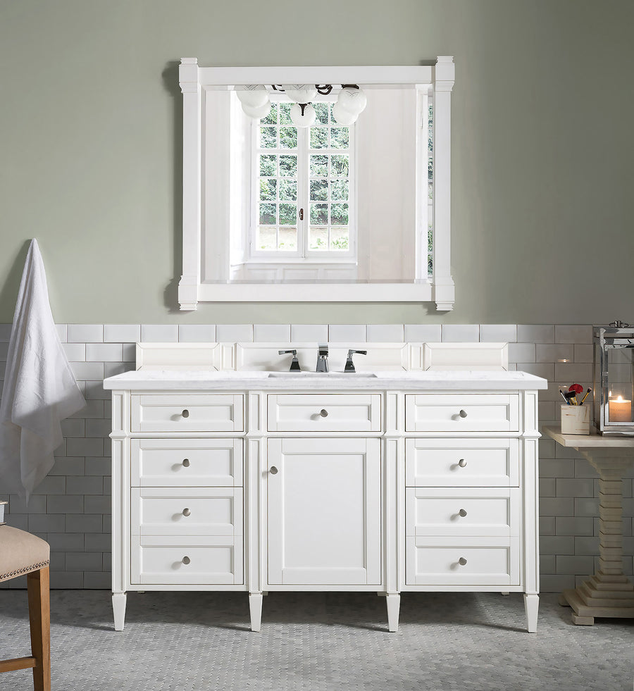 A Brittany 60" Vanity in white with multiple drawers and a large mirror is set against a light gray wall with white tiles. A towel hangs on the left, and a candle plus glass jar sit on a table to the right.
