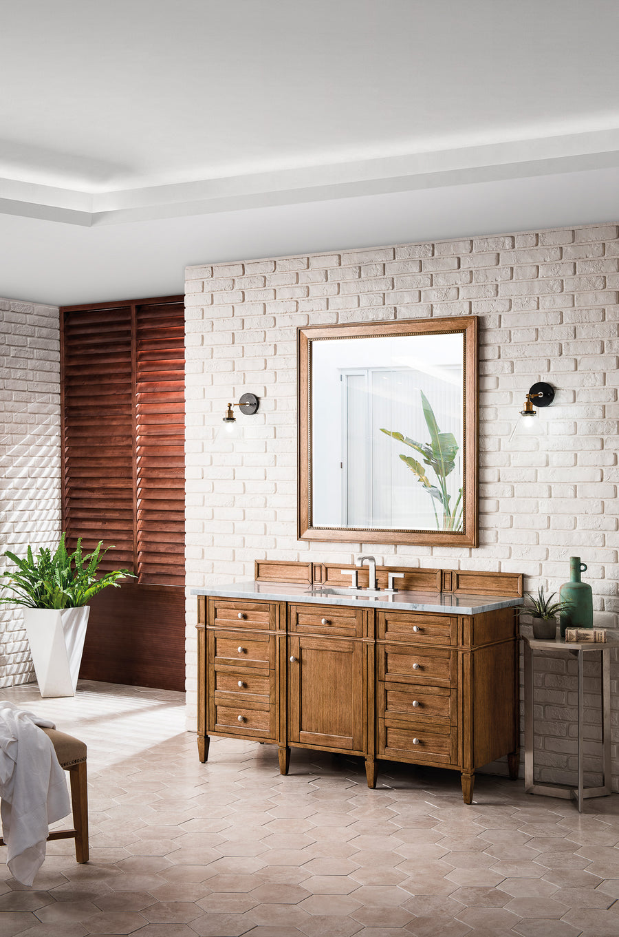 A modern bathroom with a wooden vanity and marble countertop, a large mirror, two wall sconces, potted plants, and white brick walls. Natural light enters through wooden slats in the background.