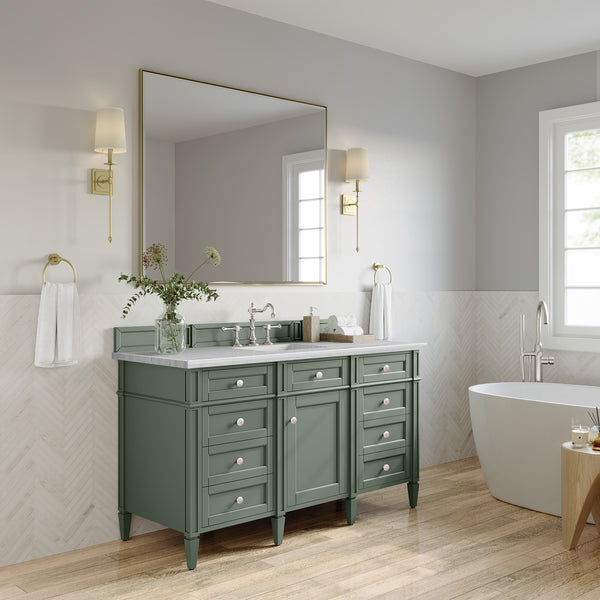 A modern bathroom with a green vanity, white countertop, large mirror, gold sconces, towel rings, freestanding bathtub, light wood floor, and chevron-patterned wall tiles. A vase with flowers sits on the vanity.