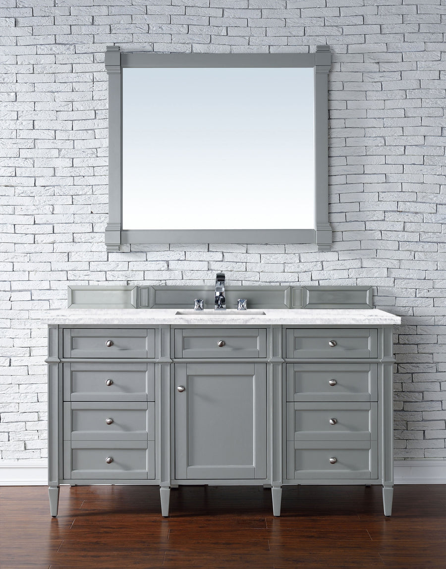 The Brittany 60" Vanity features a gray finish with a white countertop and silver handles, set against a white brick wall and topped by a matching framed mirror. It pairs beautifully with dark wood floors for a classic, timeless look.