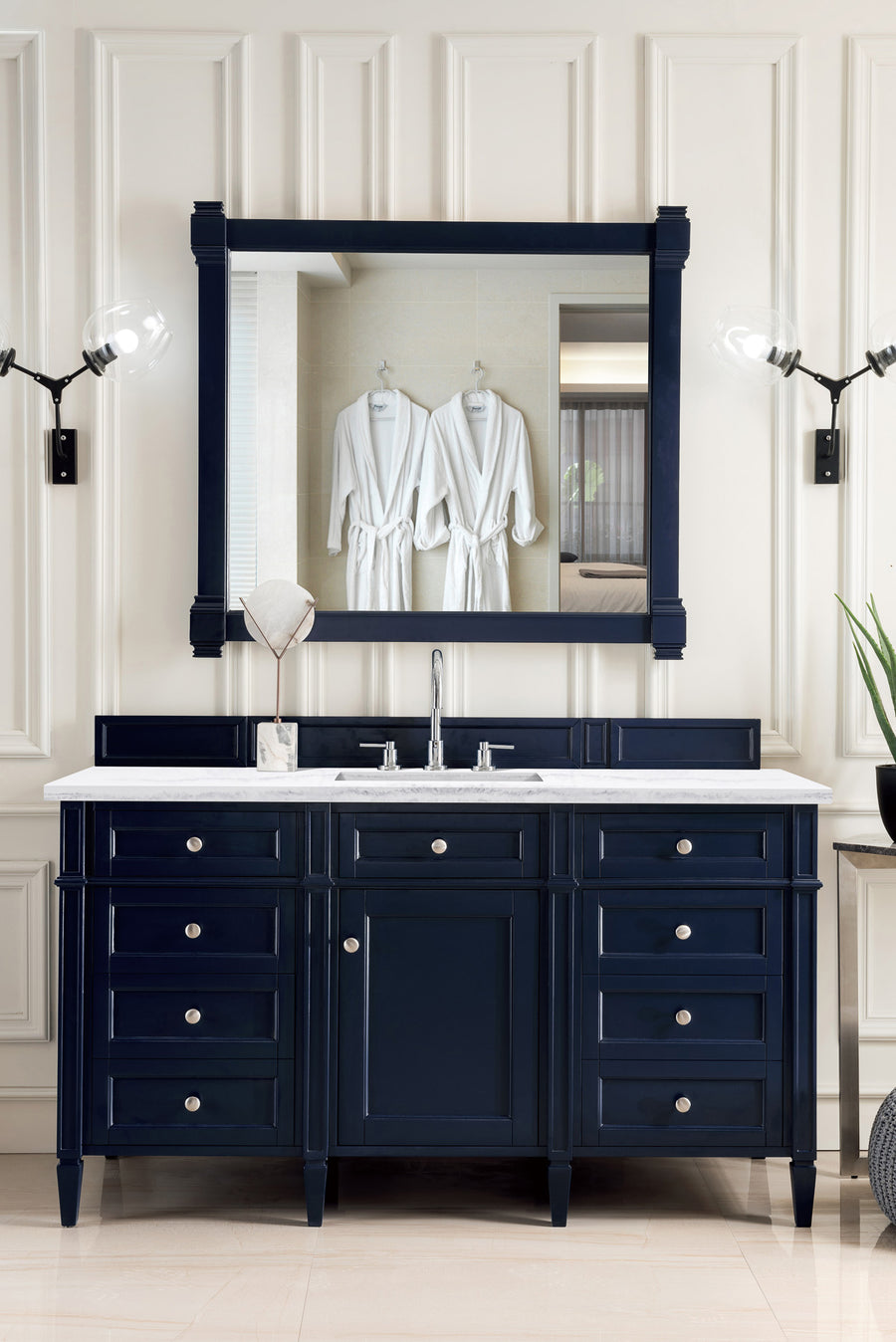 A Brittany 60" Vanity in navy blue with silver knobs and a white countertop sits below a large mirror reflecting two white robes. Two wall sconces flank the mirror, and a small plant rests on the right side of the vanity.
