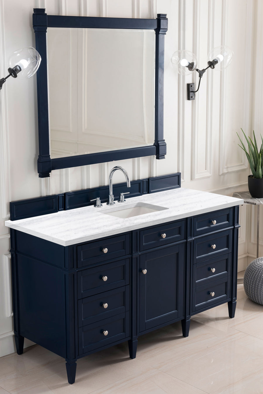 A navy blue bathroom vanity with a white marble countertop, dual sinks, and silver fixtures. A large framed mirror and two wall sconces are above, with a potted plant on the right side. The room has light-colored walls and floors.