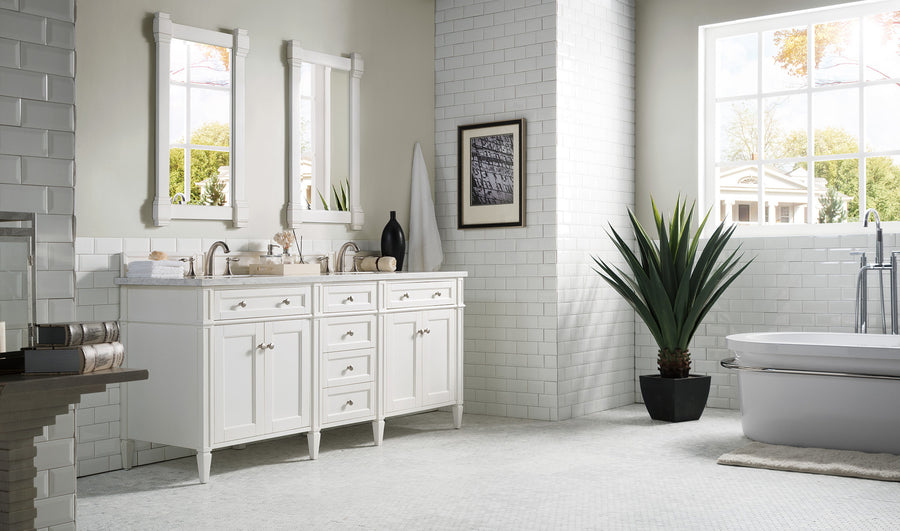 A bright, modern bathroom with white tile walls, a double sink vanity with mirrors, a potted plant, and a large window letting in natural light. A bathtub is visible on the right side of the room.
