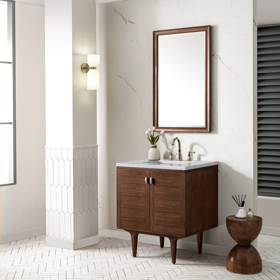 A modern bathroom features a wooden vanity with a marble countertop, a rectangular mirror with a wooden frame, brass fixtures, decorative items, and a small round wooden side table with diffusers. The walls and floor have light tile patterns.