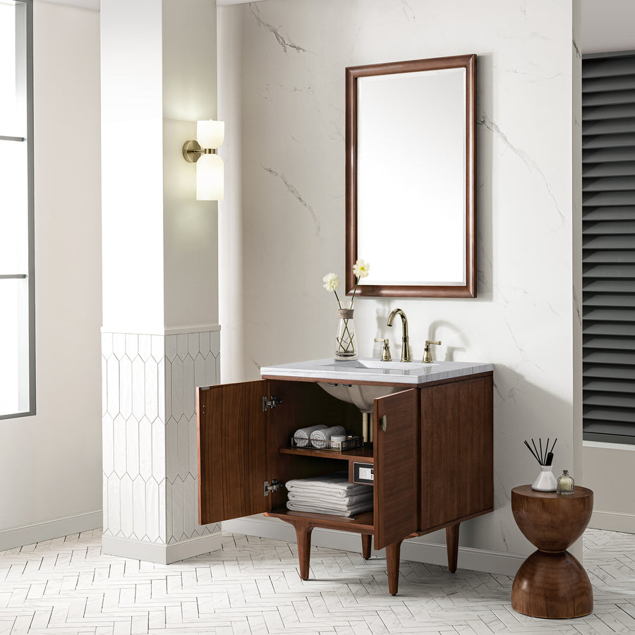 A modern bathroom with a wooden vanity cabinet, doors open to reveal shelves with towels and toiletries. A rectangular mirror, marble countertop, sink, and brass fixtures are above. Decorative items and a small wooden side table are nearby.