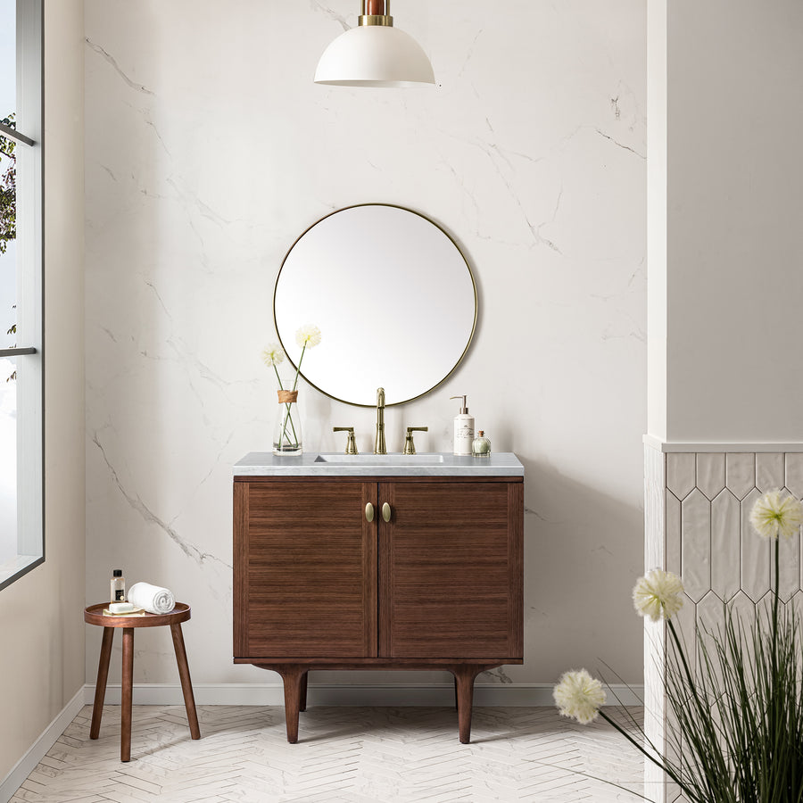 A modern bathroom featuring the Amberly 36" Vanity with a marble countertop, round mirror, gold fixtures, a small stool with rolled towel and toiletries, white walls with subtle veins, and decorative flowers.