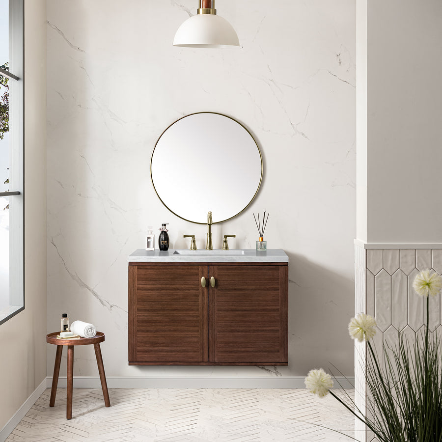 A modern bathroom features a round mirror above a wooden vanity with gold hardware, a white countertop, and gold faucet. A small wooden stool holds towels and toiletries. Minimalist decor and light walls complete the space.
