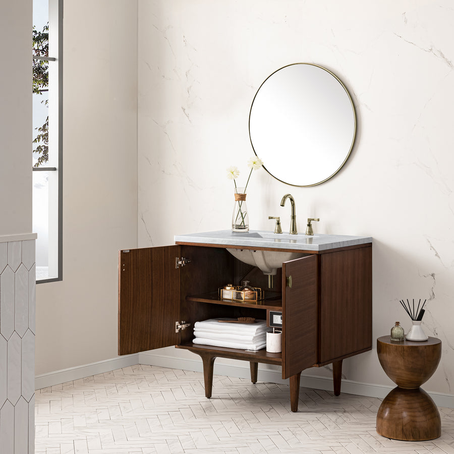 A modern bathroom with a wooden vanity, open cabinet doors showing towels and toiletries, a round mirror above, marble countertop with two vases, gold faucet, and a small wooden side table. Natural light comes from a tall window.