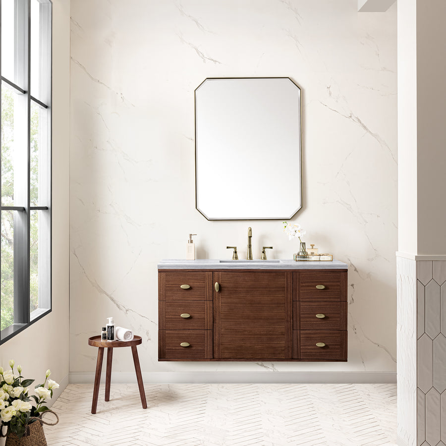 A modern bathroom with a wooden vanity, a large octagonal mirror, gold fixtures, and marble-style walls. A small round table holding towels is beside the vanity, and flowers sit in a vase on the floor by a large window.