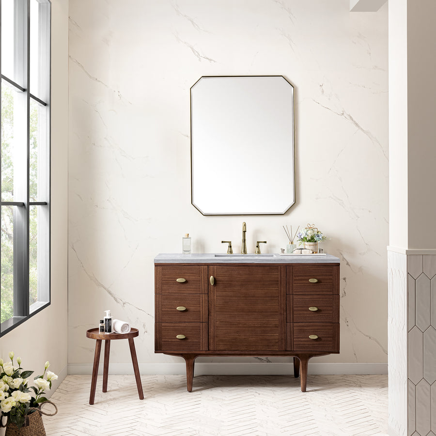 A modern bathroom featuring the Amberly 48" Vanity, brass fixtures, a large octagonal mirror, and marble-look walls. A small round table with toiletries and flowers elevates the chic décor beside a large window.