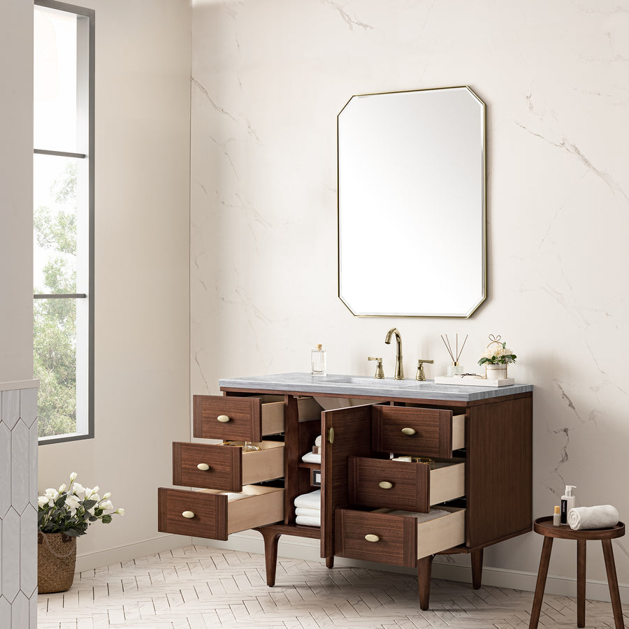 A modern bathroom with a large mirror, dark wood vanity with multiple open drawers, white marble countertop, gold faucet, and decor items. A window lets in natural light, and a small plant sits on a stool nearby.