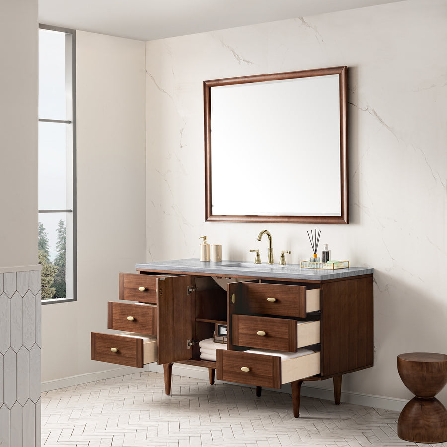 A modern bathroom with a wooden vanity featuring open drawers and doors, a marble countertop, double brass faucets, a large square mirror, and a window letting in natural light. Towels and toiletries are visible in the vanity.