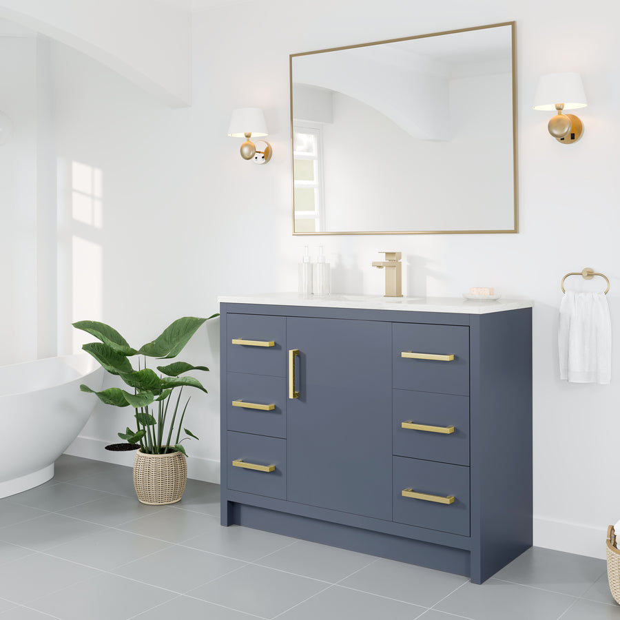 A modern bathroom with a navy blue vanity, gold hardware, a large mirror, wall sconces, a potted plant, a towel ring, and a freestanding bathtub in a bright, white space.