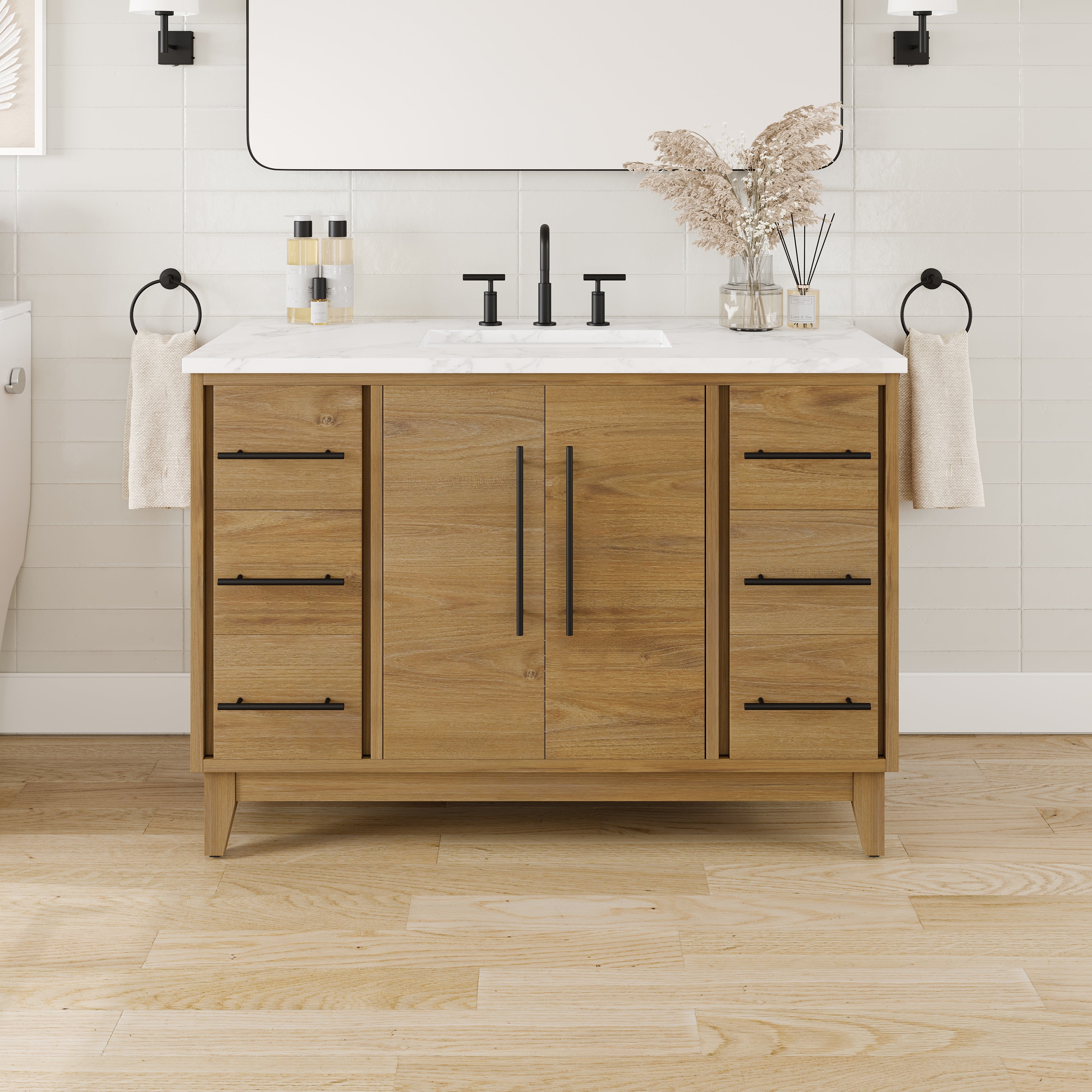 
		                            The Graham 48" Transitional Vanity in Oak features a wood finish, Arabescato engineered top, sleek black fixtures, towels on each side, decorative vases, and toiletries. Set against a white tile wall and light wood floor, this luxurious vanity combines elegance and functionality to elevate your space.
		                            