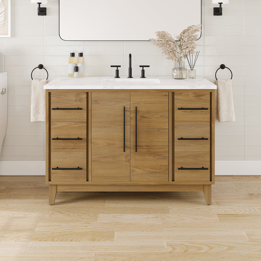 The Graham 48" Transitional Vanity in Oak features a wood finish, Arabescato engineered top, sleek black fixtures, towels on each side, decorative vases, and toiletries. Set against a white tile wall and light wood floor, this luxurious vanity combines elegance and functionality to elevate your space.