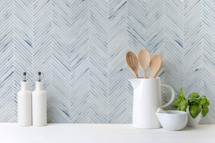 A kitchen counter features two white oil dispensers, a white pitcher with wooden spoons, a small white bowl, and a basil plant, set against a Luna Chevron Glass Tile backsplash in blue and white.