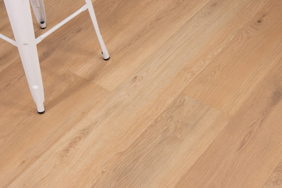 Close-up of light wood flooring with natural grain patterns. The leg of a white metal chair is visible in the top left corner of the image.